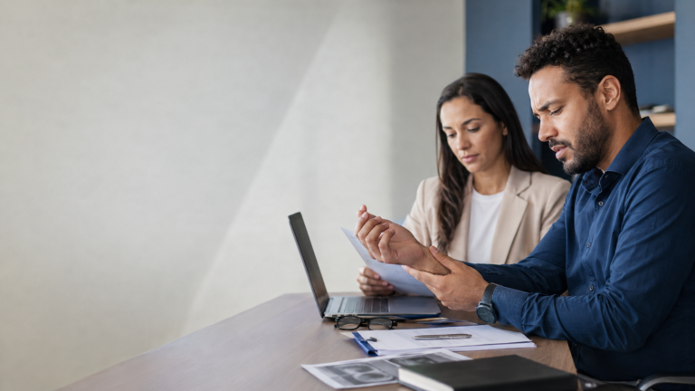 Trabalhador com desconforto no punho conversa com uma profissional enquanto analisam documentos em um ambiente corporativo.